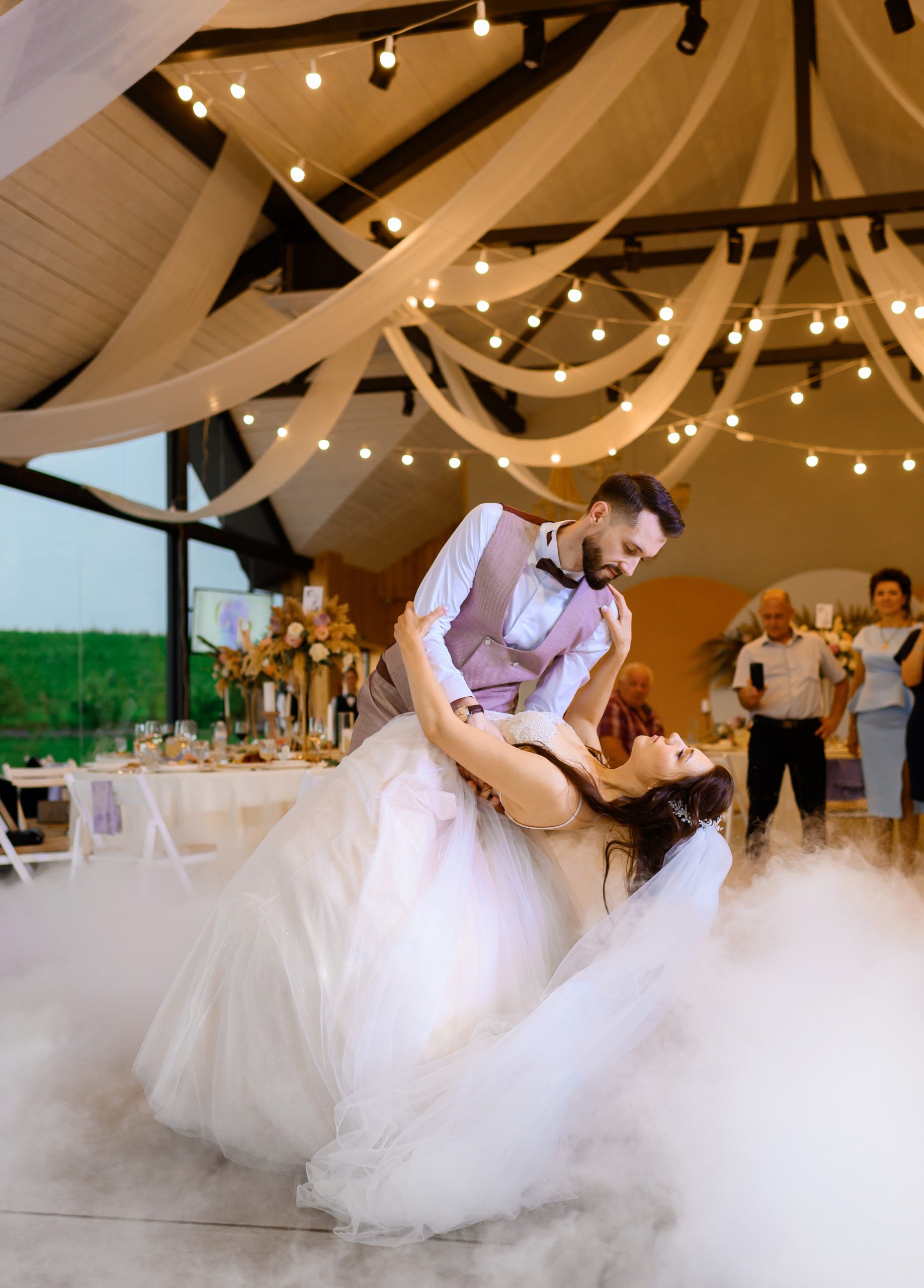 Side view of loving just married couple dancing in tent decorated with flowers, lighting garland and smoke, looking at each other while guests enjoying performance on background